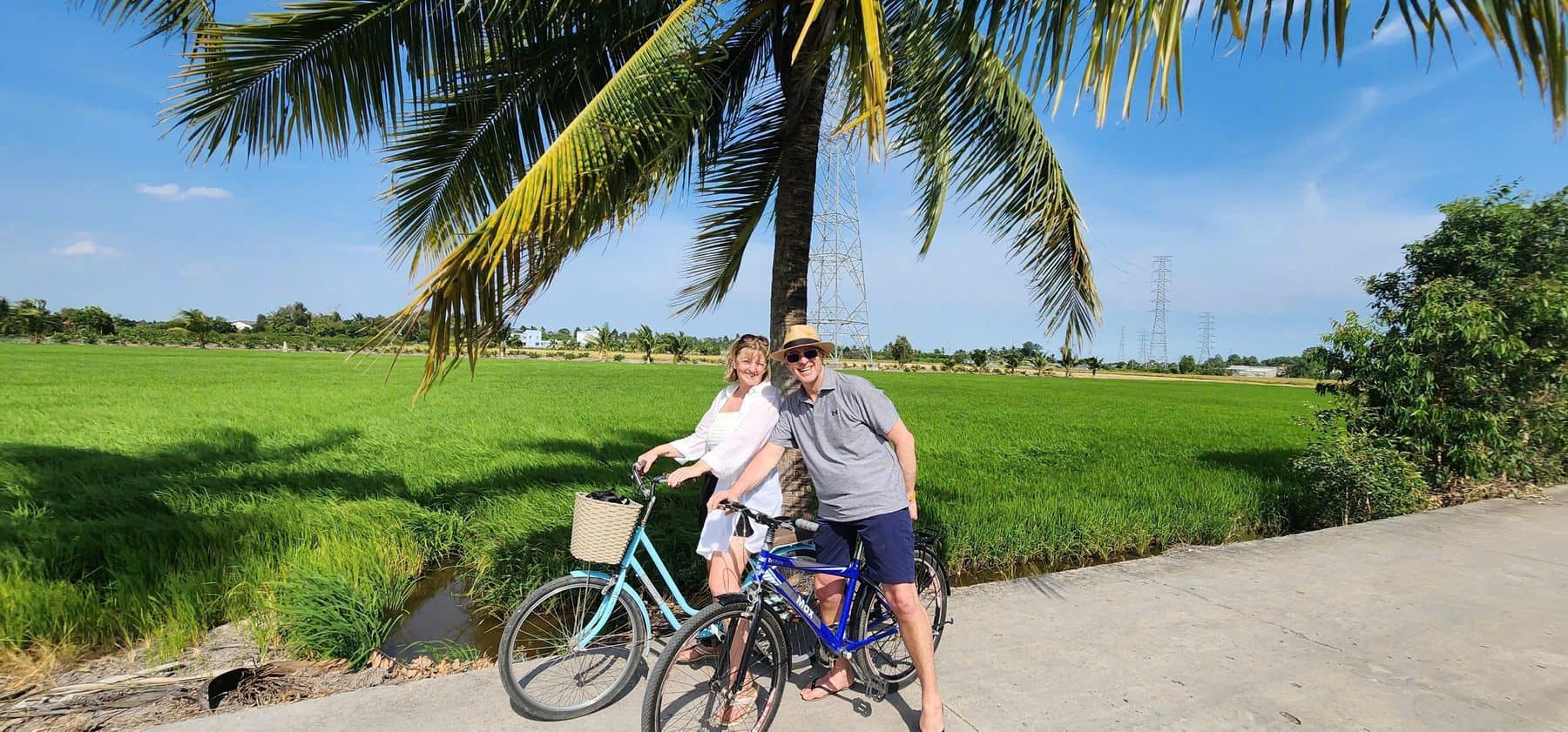 Two guests on bicycles at the edge of a rice paddy under the coconut palms