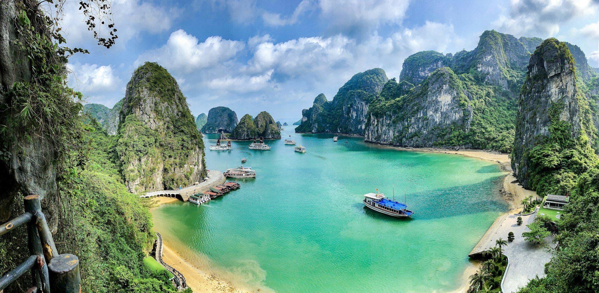 Turquoise cove ringed by limestone cliffs with tour boats and a jetty, seen from a cave path, Ha Long Bay