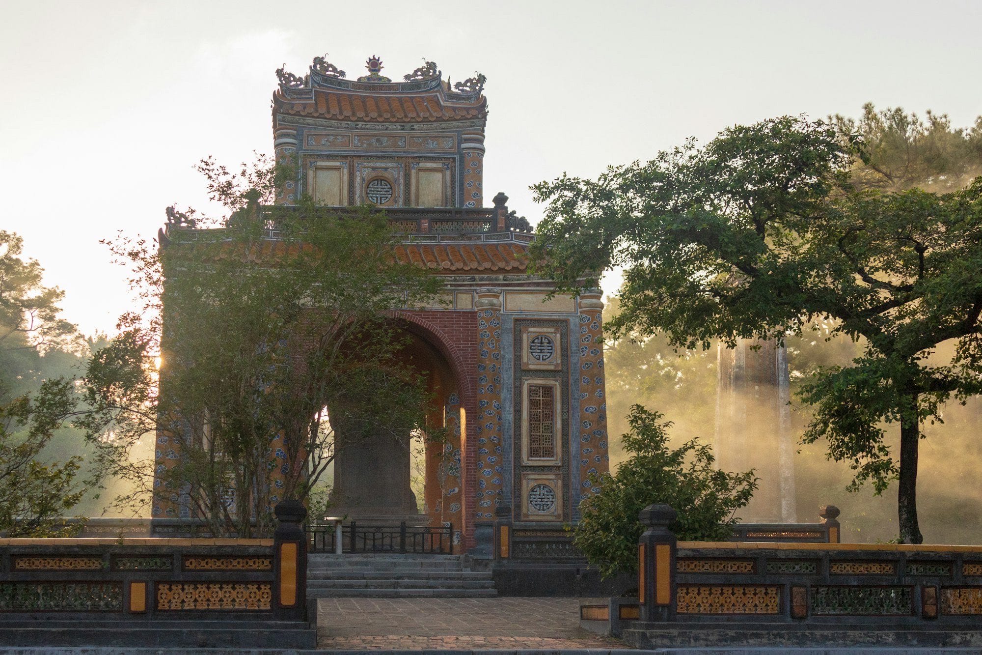 Ornate stele pavilion at Tu Duc's tomb in Hue, with sun rays filtering through misty trees