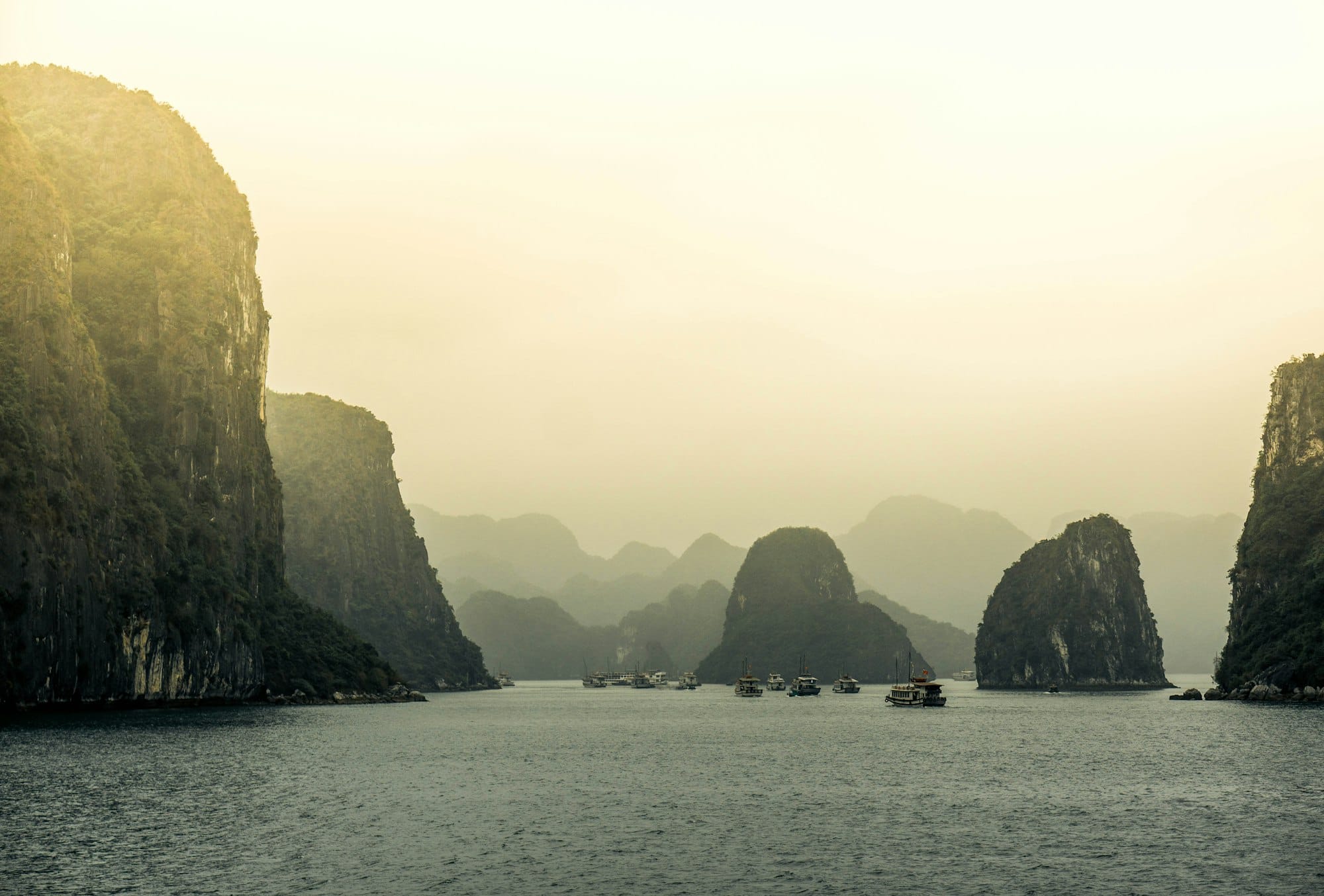 Hazy golden light over towering limestone karsts in Ha Long Bay, with cruise boats gathered on calm water