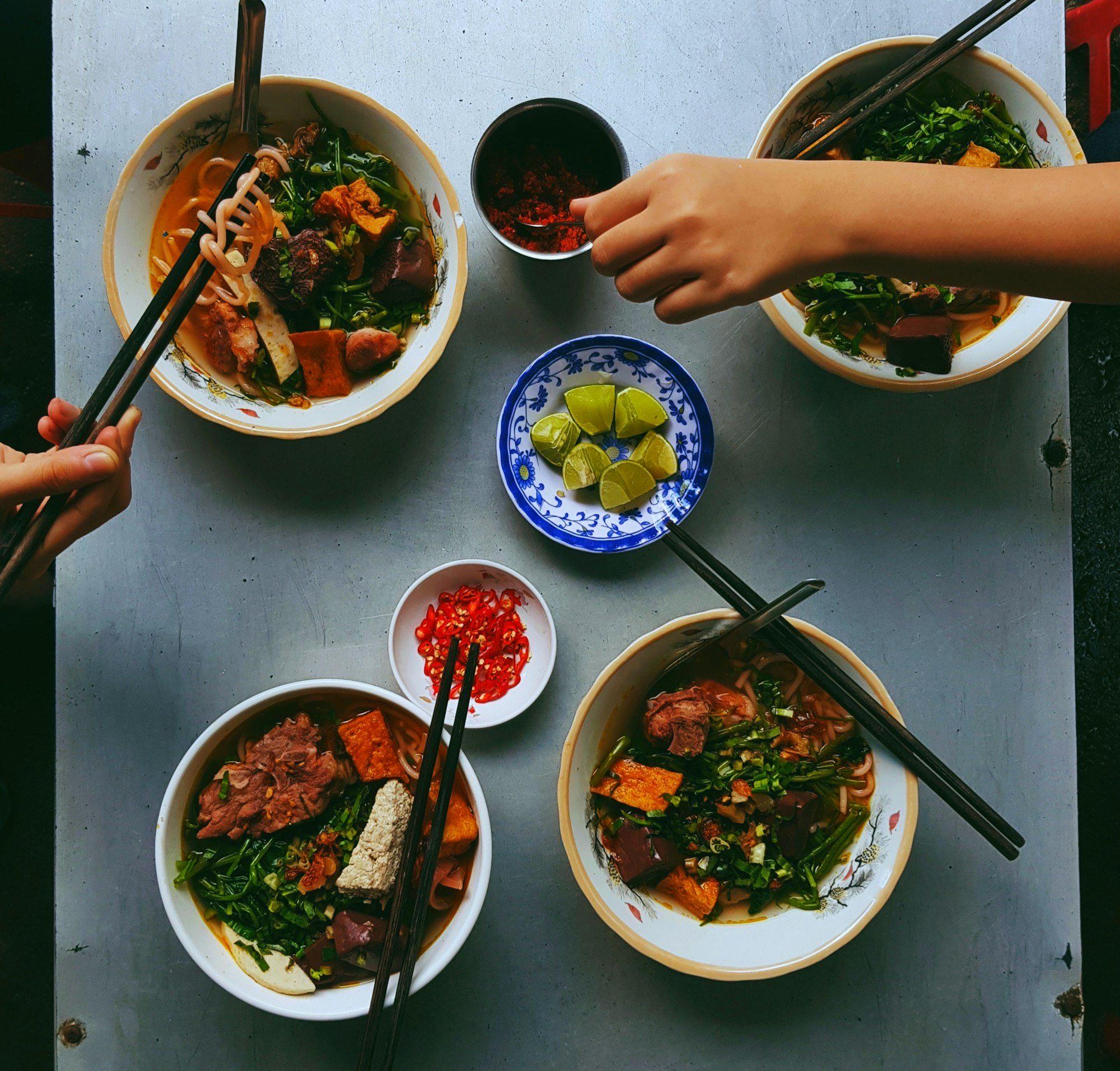 Overhead view of four bowls of bun rieu noodle soup with lime, chilli and chopsticks on a steel table