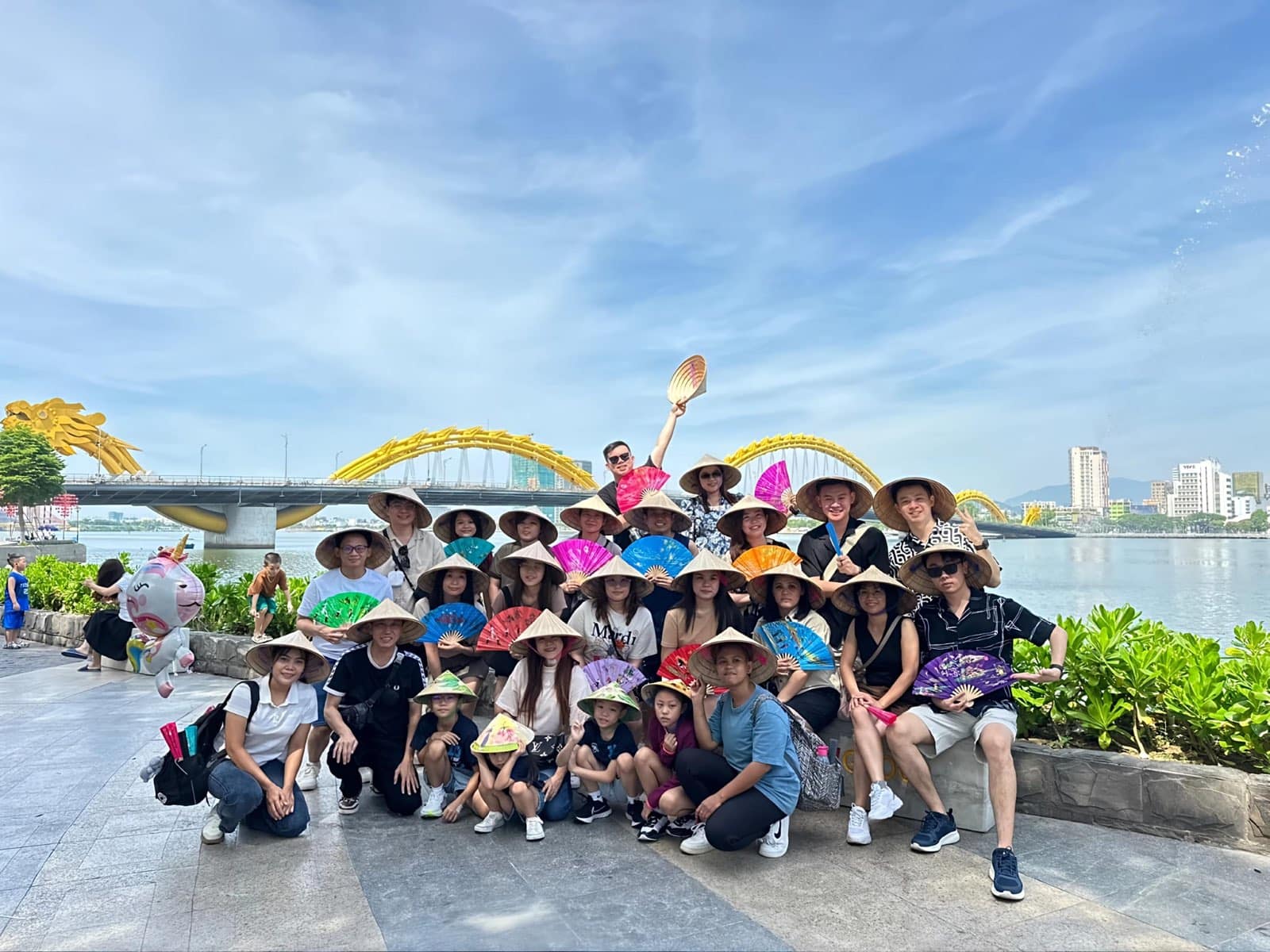 A large tour group in conical hats holding painted fans in front of the Dragon Bridge on the Han River