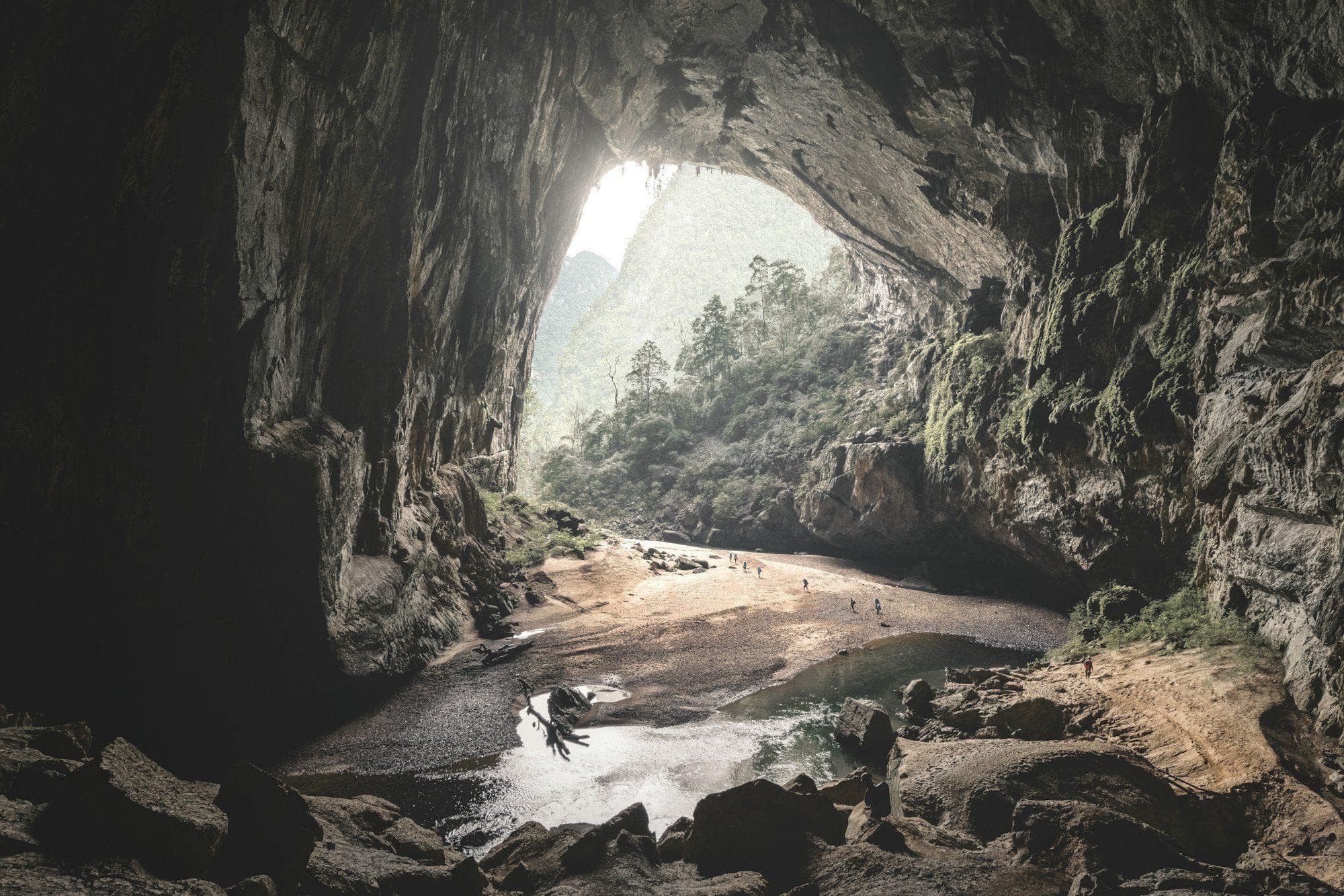 Hikers dwarfed by the vast mouth of Hang En cave as jungle light pours in, Quang Binh
