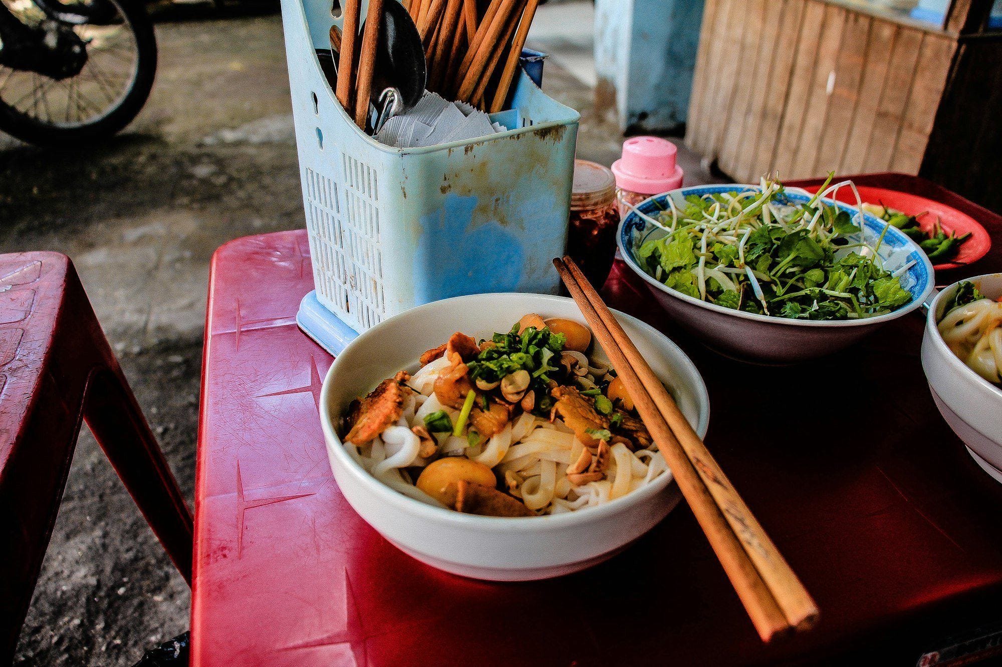 Rice noodles with pork, quail eggs and herbs on a red plastic table at a Da Nang street stall