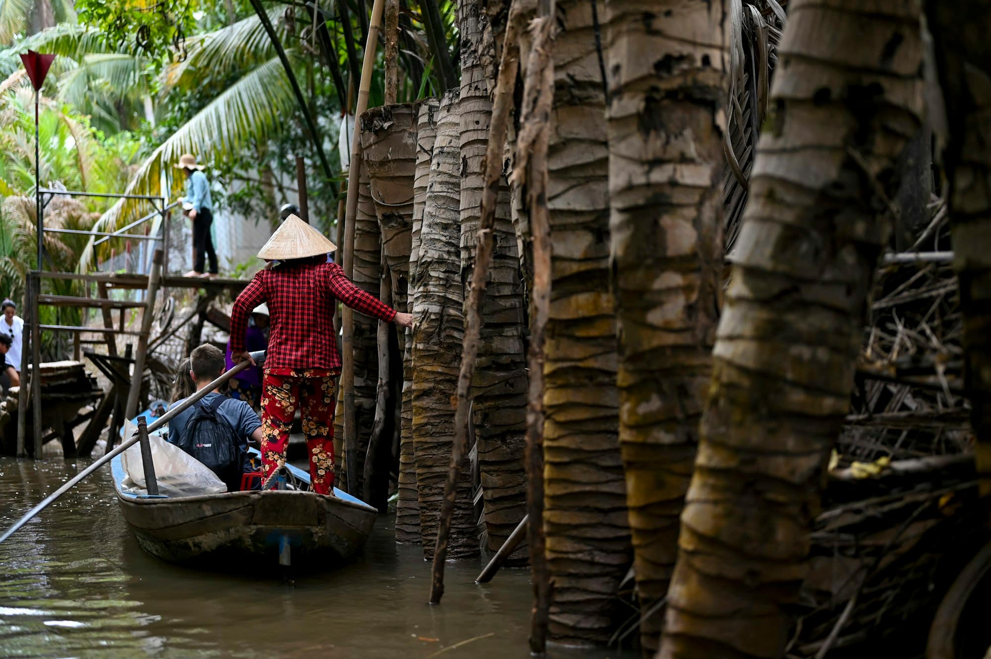 Boat woman in a conical hat poling a sampan past weathered palm trunks along a Mekong Delta canal